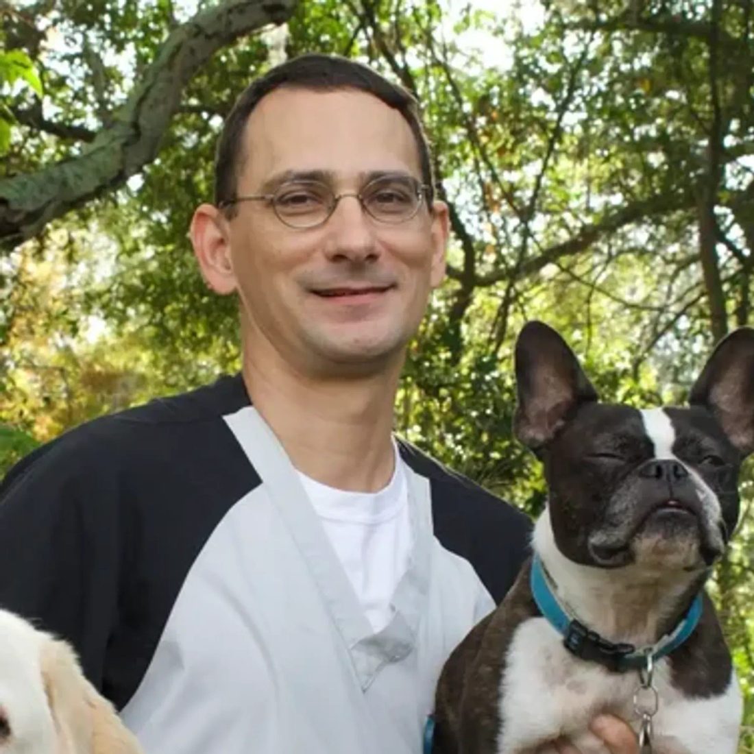 Man with glasses holding a French bulldog outdoors with trees