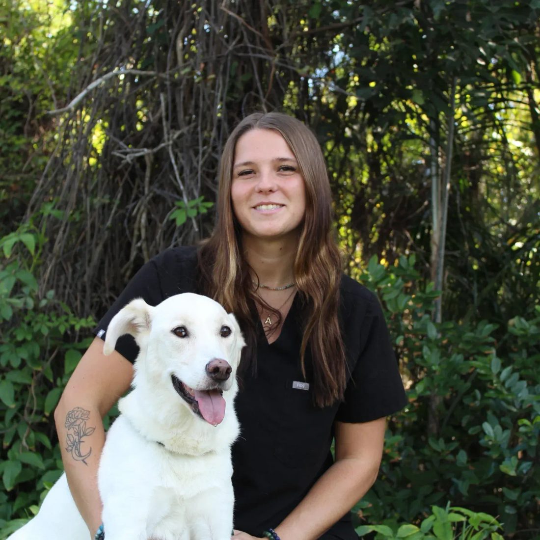 Woman in black scrubs with a white dog in front of greenery